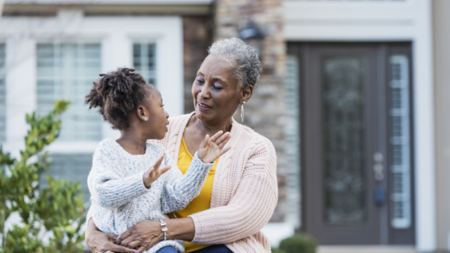 A Black grandmother and granddaughter sit outside in front of a house. The granddaughter is sitting in her grandmother's lap, looking at her grandmother, and gesticulating. The grandmother seems amused.