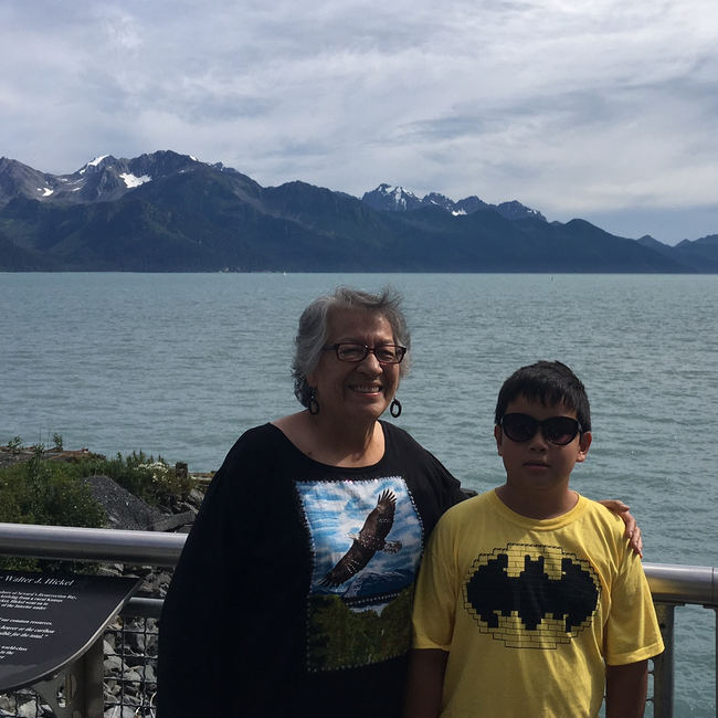 Rosalie Tallbull stands with her arm around her grandson as they pose outside in front of a beautiful vista of water and mountain