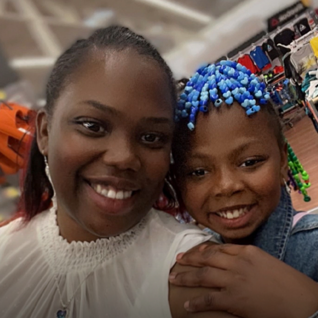 Santana Lee smiles at the camera and her daughter holds her shoulder and smiles, as well. Her daughter has blue and green beads in her hair.