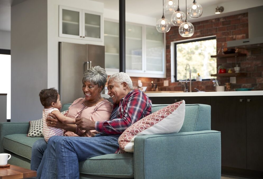 Una abuela negra sostiene a su nieto bebé, por lo que el bebé está sentado en el regazo de la abuela y frente a la abuela y el abuelo. La abuela y el abuelo sonríen mientras miran amorosamente al bebé, que los está mirando. El abuelo (que también es negro) sostiene la mano del bebé.