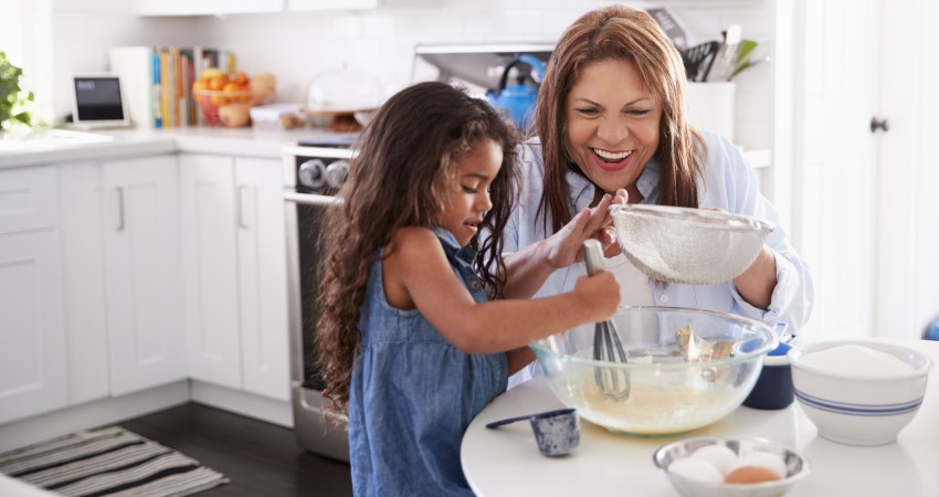 A Latina grandmother smiles as her young granddaughter uses a whisk to mix batter