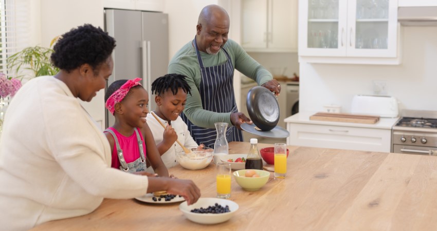 Una familia negra, compuesta por una abuela, un abuelo, una niña y un niño, sonríen mientras preparan panqueques juntos.