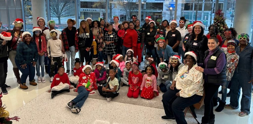 A group of families and staff members pose for a photo, with many wearing Santa hats and nametags. 