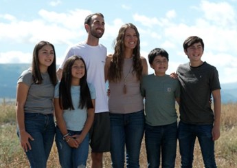 A Latino kinship family poses for a portrait outside. The adults are a young woman and a young man and there are four children/youth, two girls and two boys. 