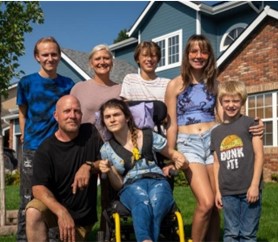 A white kin family poses for a photo in front of a house. The adults are a woman and a man, and there are five children/youth, two girls and three boys. One of the girls has a visible disability.