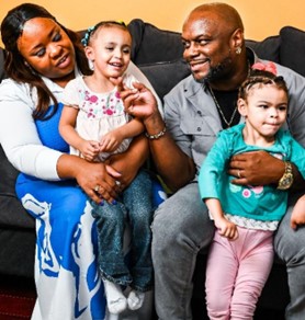 A Black kinship family sits together on a couch. The two adults are a woman and a man. The woman has a little girl in her lap and the man is holding another little girl.