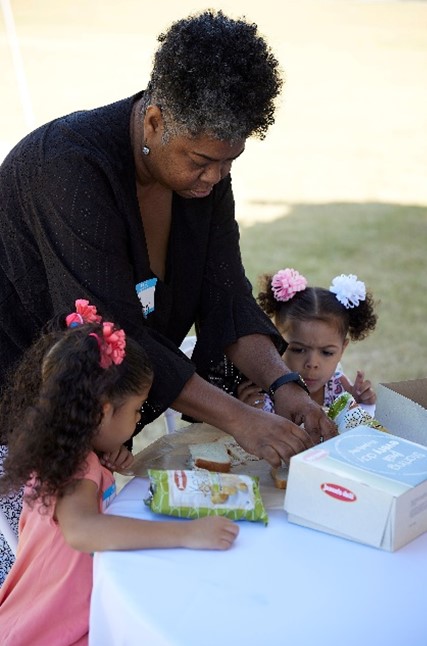 An African American grandmother helps her two young granddaughters open their food at an outdoor event
