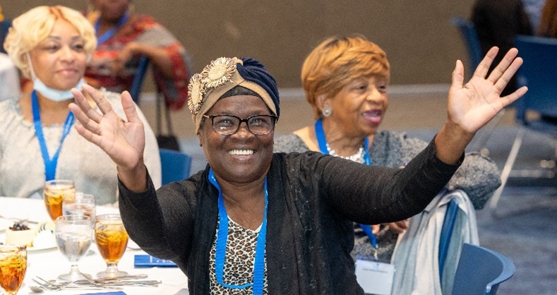 Three African American women sit together at a banquet. One is facing the camera and smiling widely with her arms in the air.