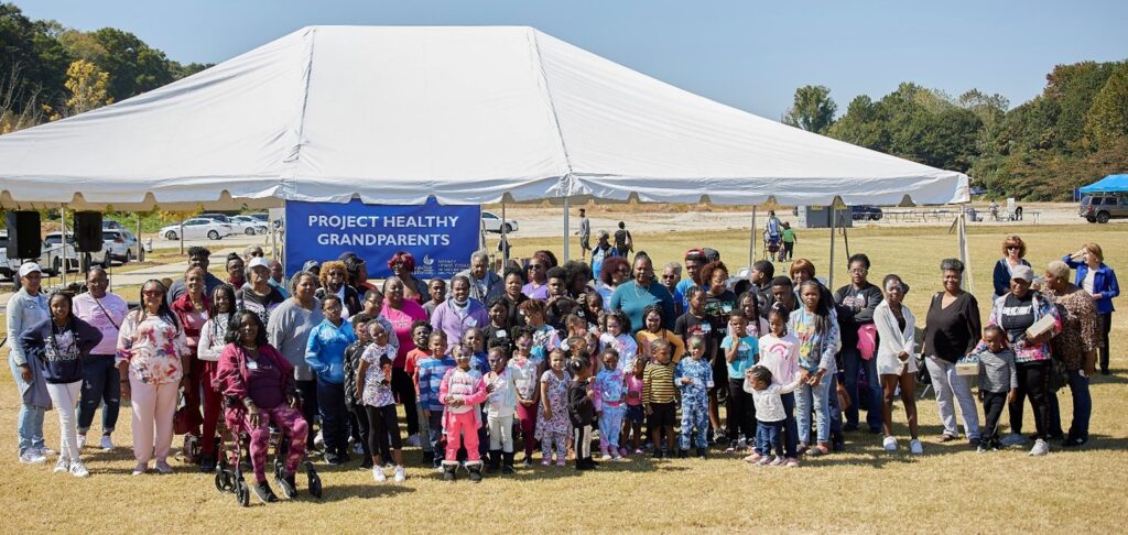 A large group of people, adults and children, nearly all African American, pose in front of a large white tent and a sign that says PROJECT HEALTHY GRANDPARENTS