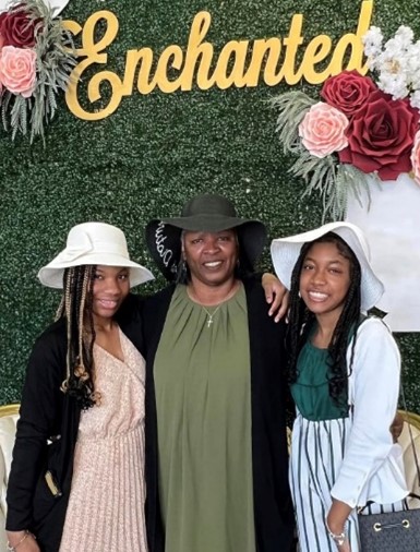 A kin caregiver and the two children in her care, both tween or teenage girls, pose for a photo under the word "Enchanted," with decorative flowers on the wall behind them. All three are wearing fancy hats.