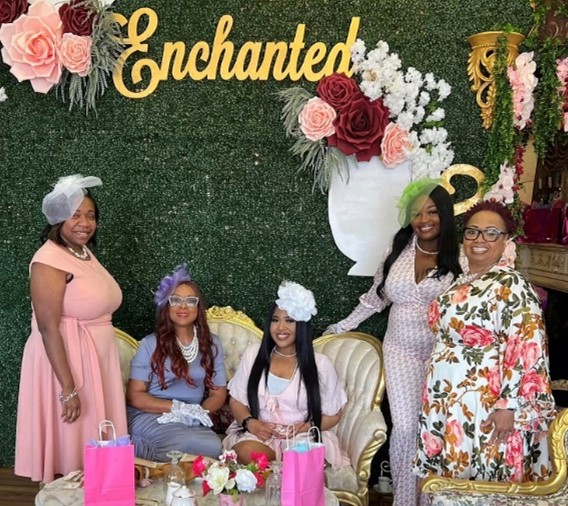 A group of women pose for a photo under the word "Enchanted," with decorative flowers on the wall behind them and gift bags on the table in front of them. 
