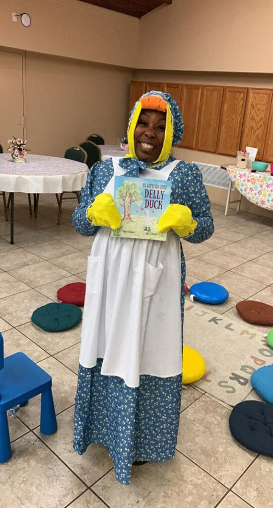 A smiling Black woman, dressed as a duck wearing a long dress and apron, holds up a picture book