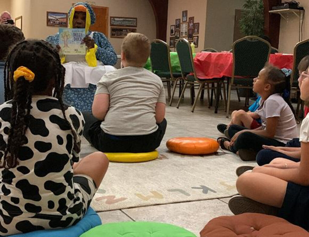 A small, diverse group of children sits on cushions on the floor as a Black woman, dressed as a duck wearing a long dress and apron, sits on a chair in front of them and holds up a picture book