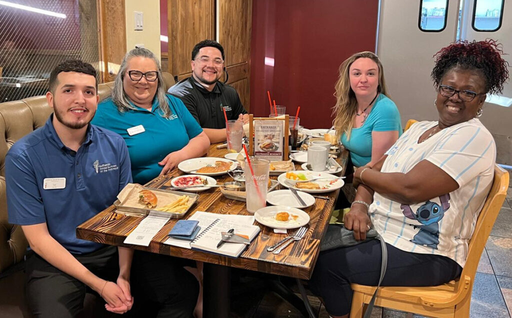 A diverse group of five adults sits at a restaurant table together and smiles for a picture