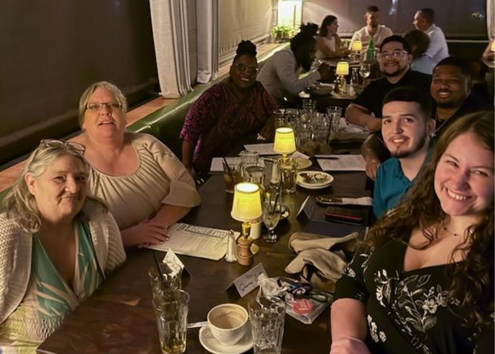 A large, diverse group of adults sits at a long restaurant table and smiles for a picture