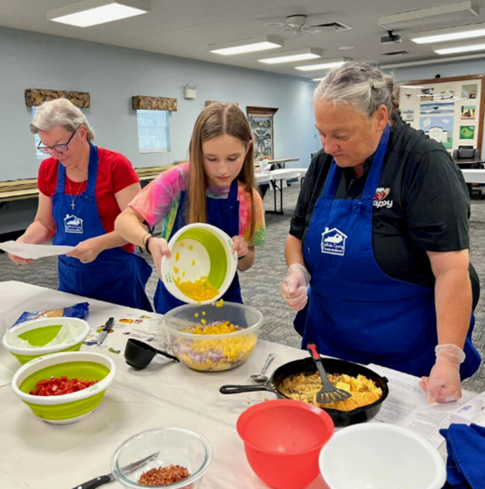 Two older adult women and a girl, all white and wearing t-shirts and blue aprons, stand behind a table and prepare food together