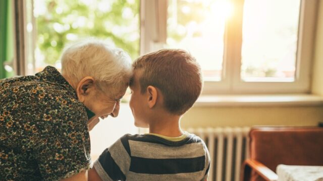 An older white woman smiles and looks at her elementary-aged great-grandson and leans her head against his head. Both individuals have their backs to the camera, but the great-grandmother’s head is in profile and the great-grandson’s head is turned slightly toward his great-grandmother.