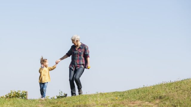 A young girl and her grandmother hold hands and walk down a hill in a grassy field. Both individuals are white.