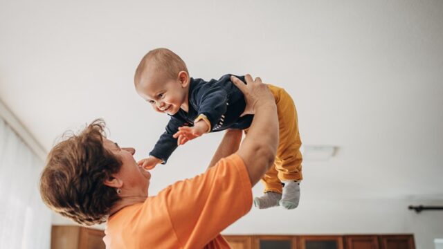 A smiling white grandmother looks at her baby grandson as she holds him in the air above her head. The grandson smiles and reaches his hands out towards his grandmother.