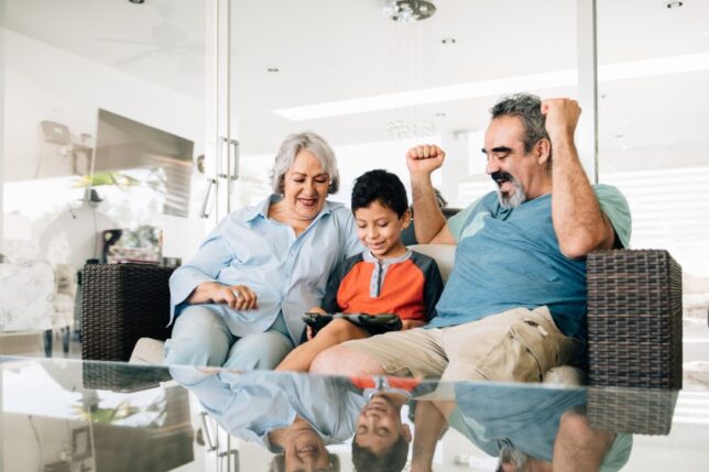 A Hispanic/Latino grandmother, grandson, and grandfather play a video game together on the couch, and the grandfather pumps his arms in celebration.