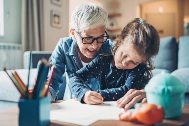A white grandmother smiles and leans over her young granddaughter's shoulder as the child writes/colors
