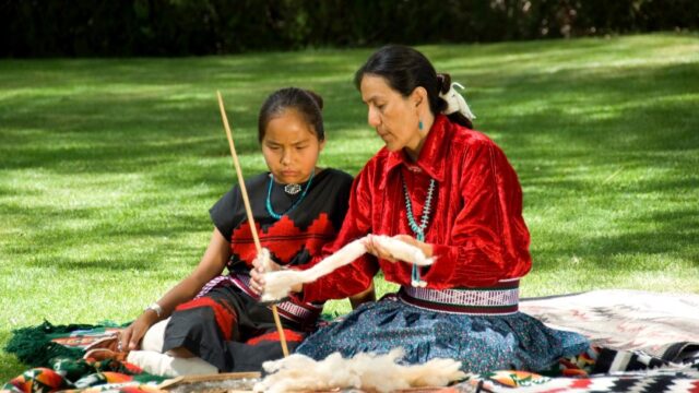 A Navajo woman and child sit outside on a blanket and the woman shows the child how to weave