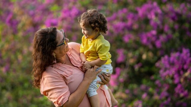 A Hispanic/Latina woman holds her young grandchild outside and the two smile at each other. Behind them is a bush in bloom.