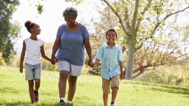 A Black grandmother holds hands with her granddaughter and grandson and walks across a field. The grandmother and granddaughter are looking at each other and smiling. The grandson is looking forward and smiling.