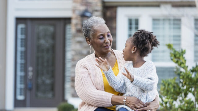 A Black grandmother and granddaughter sit outside in front of a house. The granddaughter is sitting in her grandmother's lap, looking at her grandmother, and gesticulating. The grandmother seems amused.