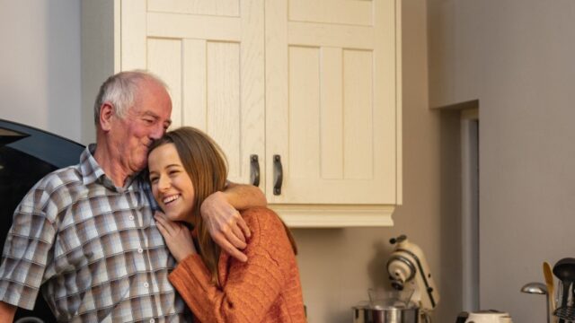 A smiling teenage girl and her grandfather stand in a kitchen together. The grandfather has his arm around the girl’s shoulders and is kissing the top of her head, and the granddaughter has her hand on her grandfather’s chest. Both individuals are white.