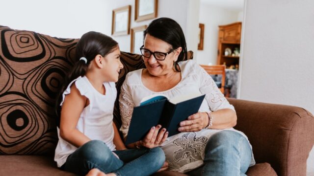 A Latina grandmother and granddaughter look at each other as they read a book on the couch together