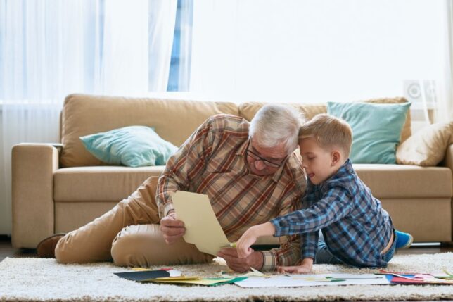 A white grandfather and young grandson sit on the floor and look at papers together.