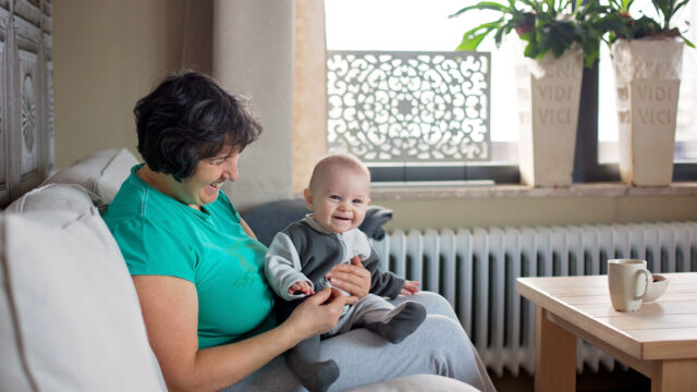 A white grandmother sits on a couch a smiles as she holds her smiling baby grandchild.