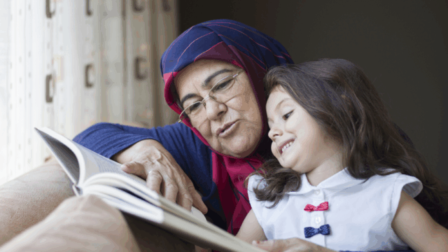A grandmother in a headscarf reads with her young granddaughter, who is smiling