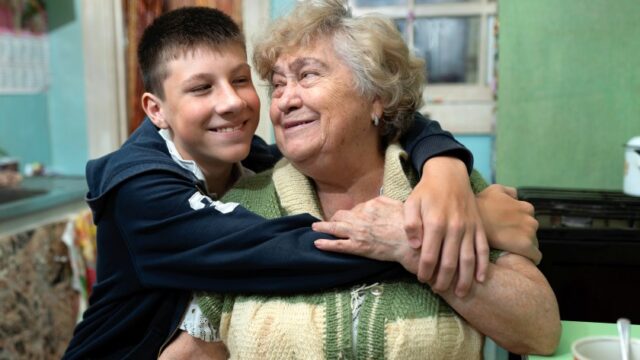 A white grandmother and teenage grandson smile at each other as they sit together. The grandson is hugging his grandmother, and she has her hand on his arm.