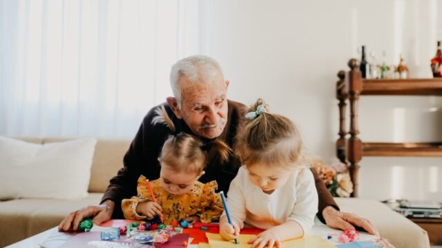 Grandfather sitting behind two grandchildren that are drawing on colored paper on a table.