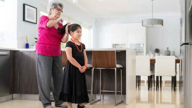 A girl in a fancy dress smiles as her grandmother does her hair. The family is Cuban-American.