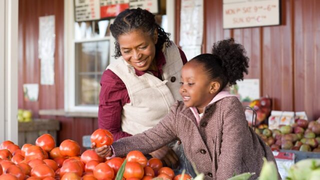 A Black grandmother and granddaughter smile as they pick out fruits at a farm stand