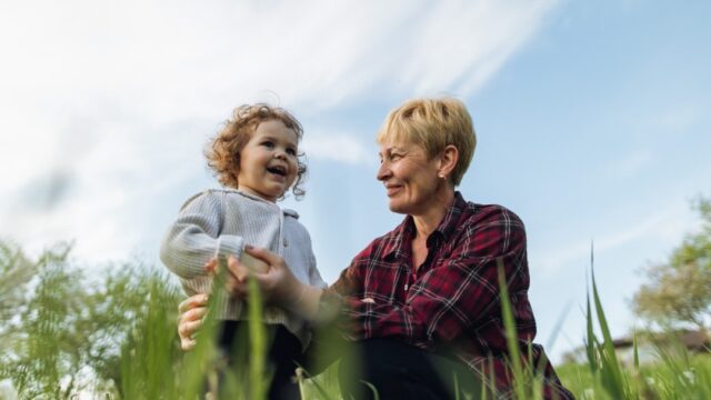 A white grandmother and her young grandchild stand in a field together. The grandmother is sitting or kneeling and looking at and holding onto her grandchild and the child is smiling.