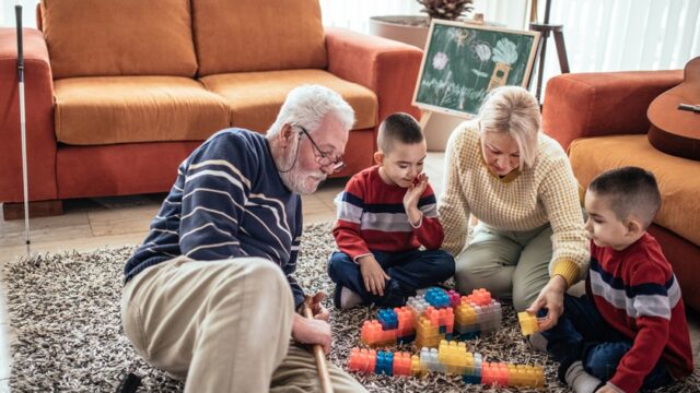 A white grandmother and grandfather sit on the floor with their twin grandsons, playing with large, Lego-type blocks