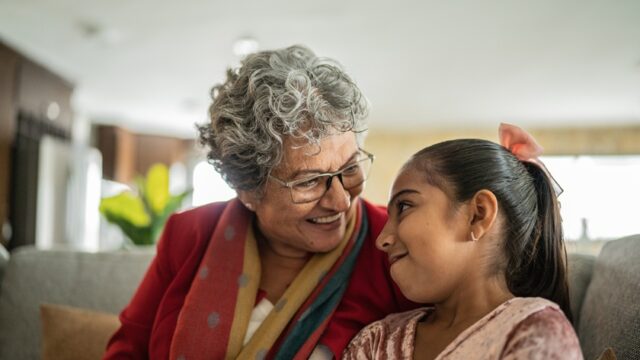 A Mexican grandmother and granddaughter smile as they look at each other and lean into each other while sitting on the couch together
