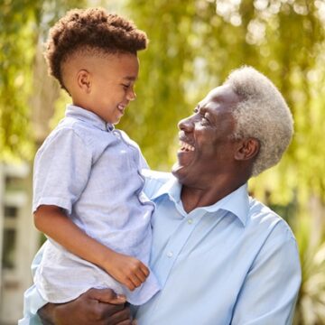A Black grandfather holds his grandson and the two of them look at each other and smile