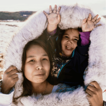 A young Inuit woman holds a girl in her agouti or parka hood. Both are facing the camera and smiling, and the girl has a large smile and her hands in the air. They are on the tundra.