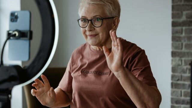 An older woman gestures as she films herself using a smartphone and a ring light