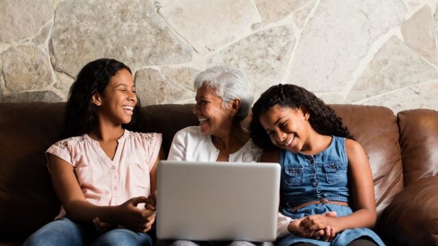 A Latina grandmother and her two tween and teen granddaughters smile as they sit on a couch together with a laptop