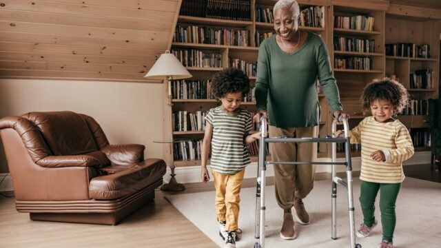 An African American grandmother smiles as she walks with her young grandchildren, who are both holding her walker along with her