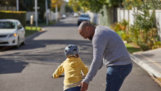 An African American man smiles as he holds the seat of a bicycle and helps a young boy learn how to ride down the street