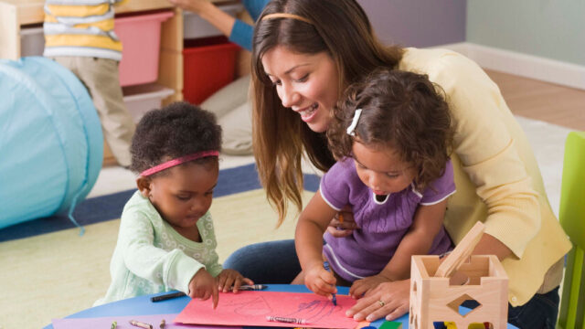 A young child care worker smiles as she watches two toddler girls draw together. One child is in her lap and the other is standing next to the table. In the background, another adult and child are slightly visible.