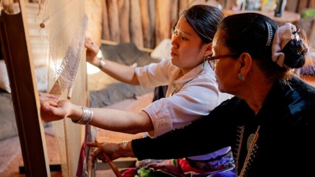 A Native grandmother helps her granddaughter use a loom to weave a traditional design