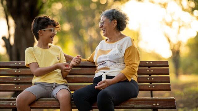 A Latina grandmother and her grandson, both wearing glasses, sit on a park bench and smile as they look at and talk with each other
